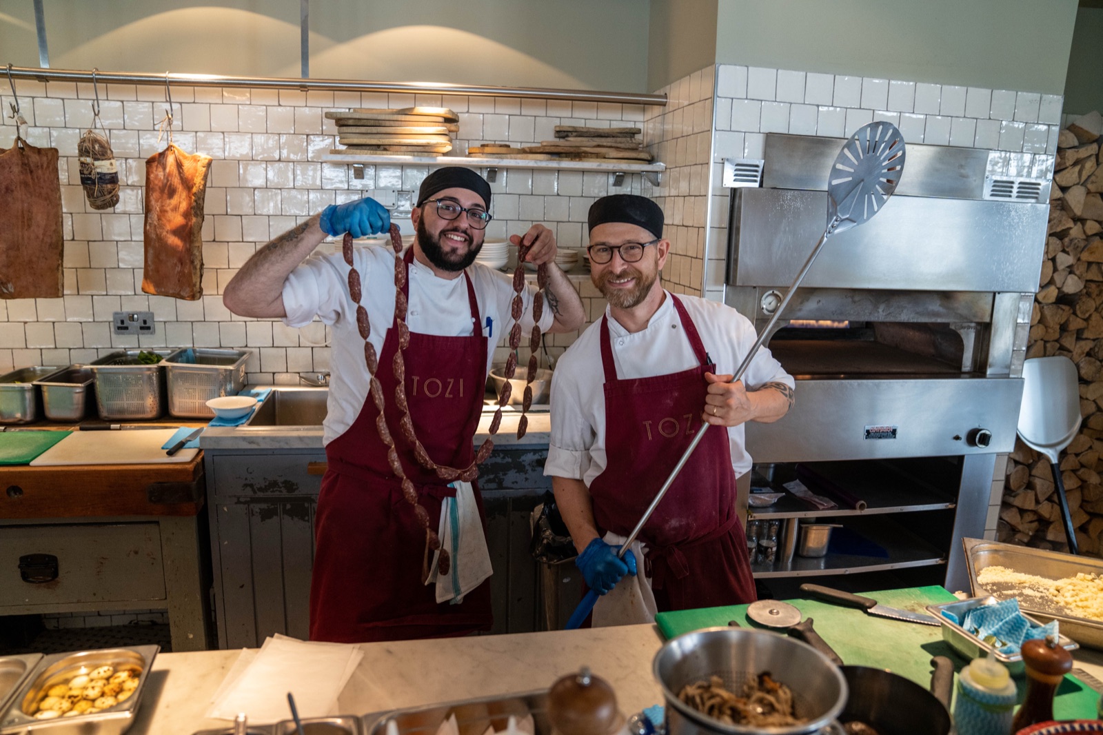 Two chefs in TOZI aprons holding fresh sausage and a pizza peel in the restaurant's open kitchen